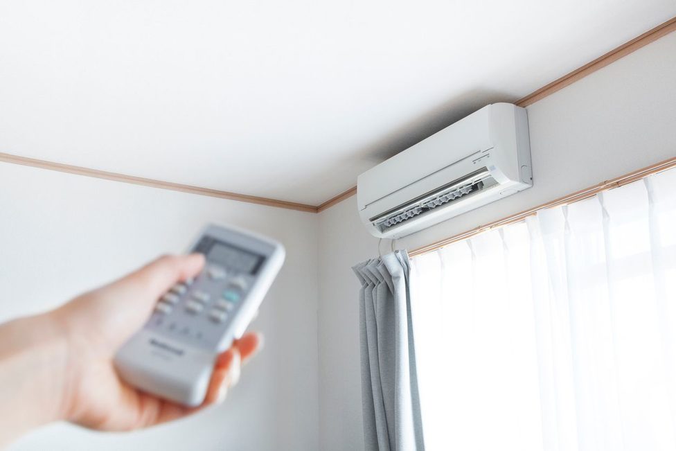 Una mano que sostiene un control remoto apunta hacia una unidad de aire acondicionado montada en la pared instalada cerca del techo junto a una ventana con cortinas blancas.