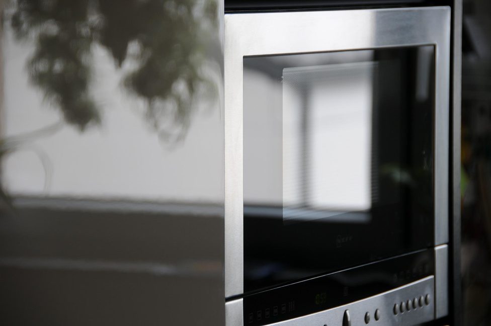 Un microondas de acero inoxidable con la puerta entreabierta, que refleja detalles borrosos de una habitación y plantas de interior.