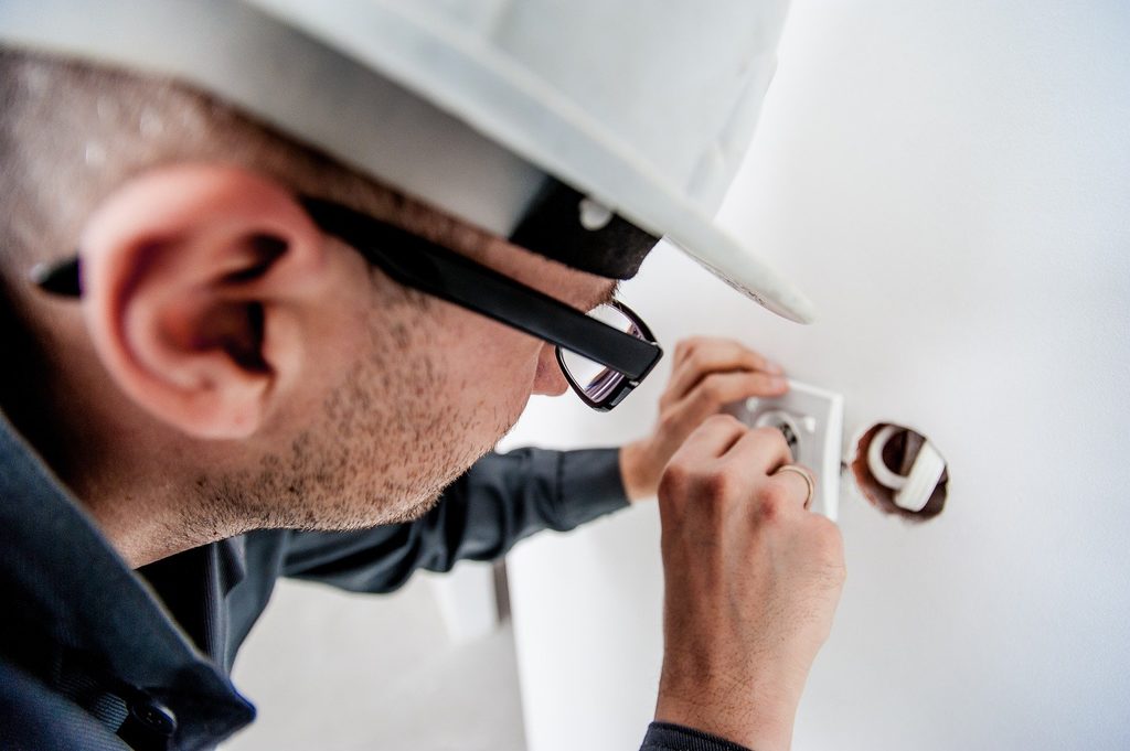 Una persona que lleva casco y gafas instala o repara una toma de corriente en una pared blanca.