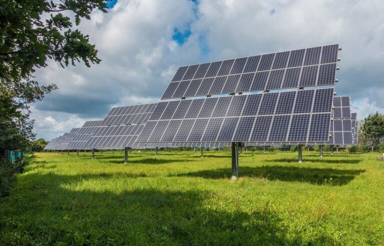 Un campo con múltiples paneles solares montados sobre soportes metálicos, frente a un cielo parcialmente nublado. Los árboles son visibles al fondo.