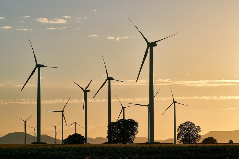 Un campo de turbinas eólicas al atardecer con árboles dispersos alrededor y colinas al fondo.