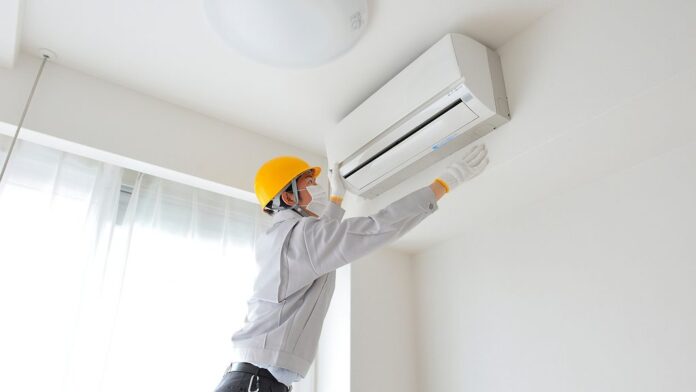 portada-clima-2000px_0 Un técnico que lleva casco, máscara y guantes está inspeccionando una unidad de aire acondicionado montada en una pared en una habitación luminosa con grandes ventanas.