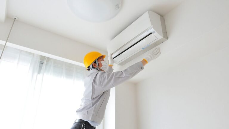 Un técnico que lleva casco, máscara y guantes está inspeccionando una unidad de aire acondicionado montada en una pared en una habitación luminosa con grandes ventanas.