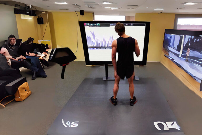 Una persona en ropa deportiva se para sobre una colchoneta e interactúa con una pantalla grande en una habitación moderna y bien iluminada, mientras que otras dos se sientan en un sofá cercano, mirando tabletas.