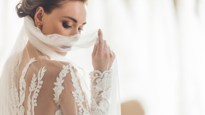 SendPyme- Atelier de novias Una novia con un vestido de novia de encaje sostiene suavemente su velo, mirando hacia abajo.