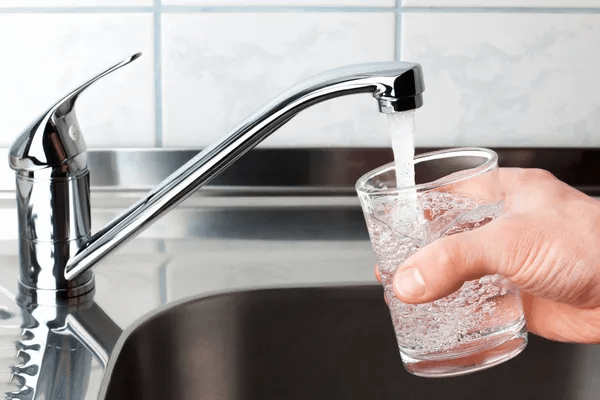 Una persona llena un vaso transparente con agua de un grifo de cocina de metal sobre un fregadero.